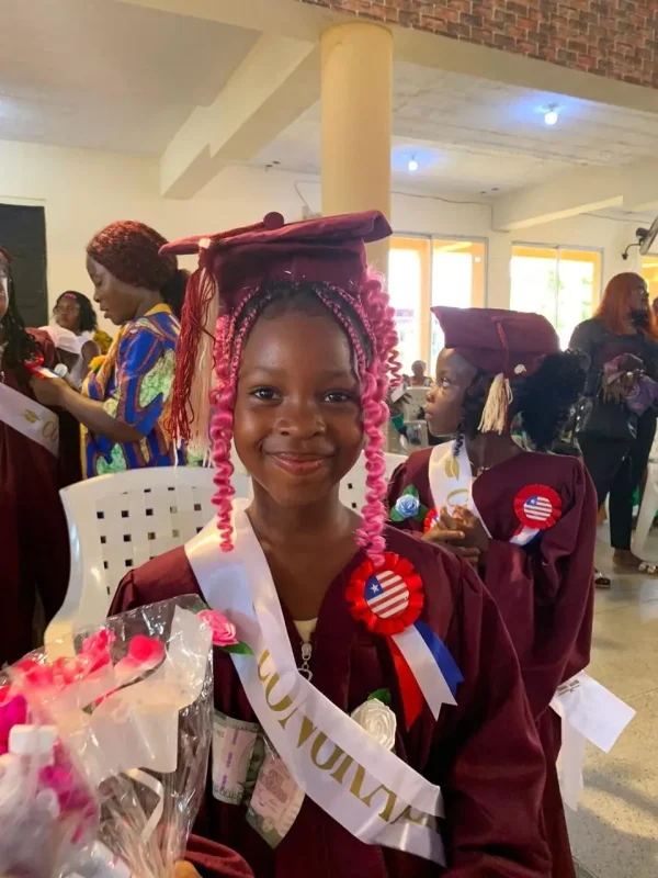 A Zoe Robinson Academy kindergarten graduate in a maroon cap and gown, smiling and holding a bouquet after the ceremony.